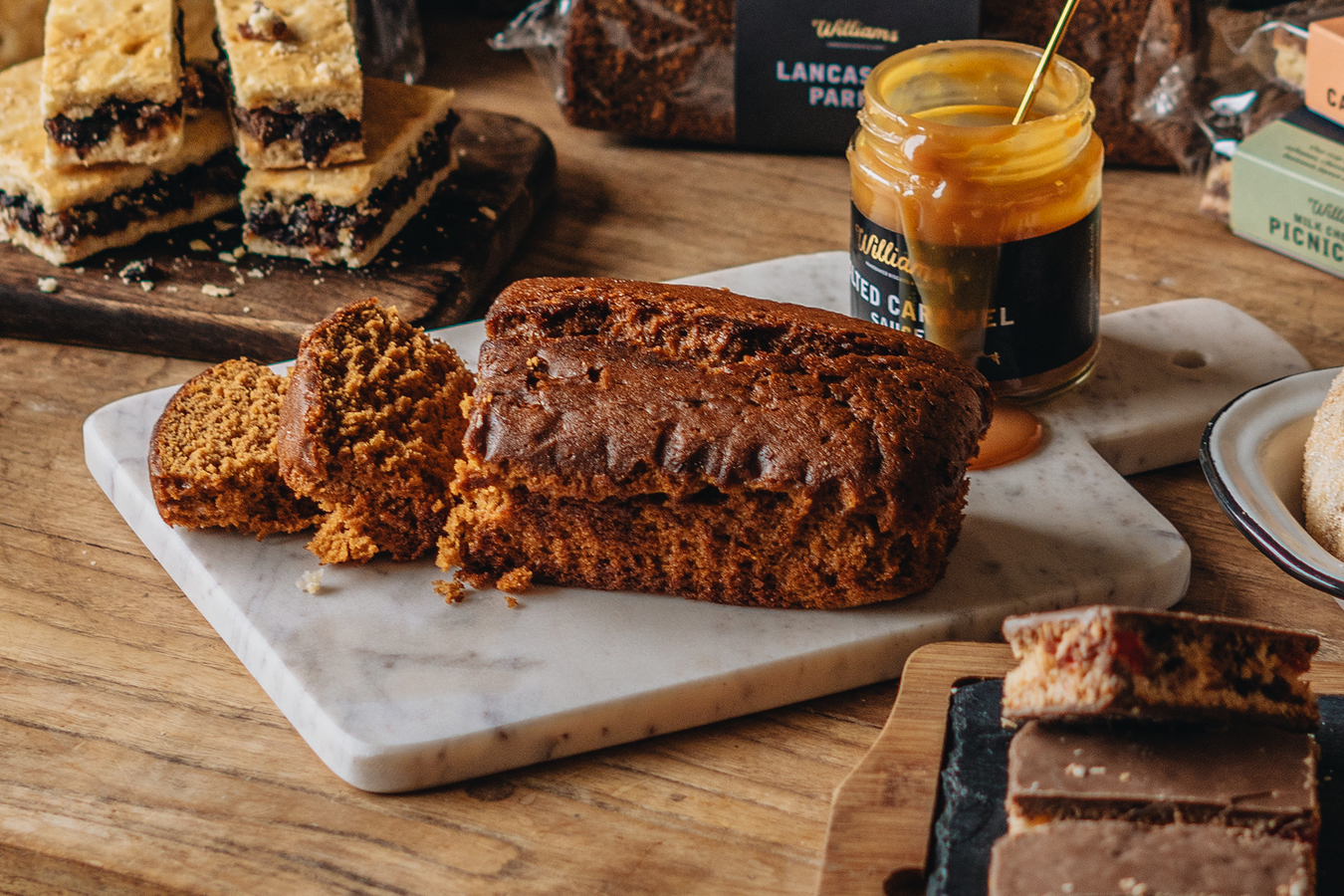 A loaf cake sliced up on a marble serving board with an open jar of salted caramel sauce behind, surrounded by other bakes.
