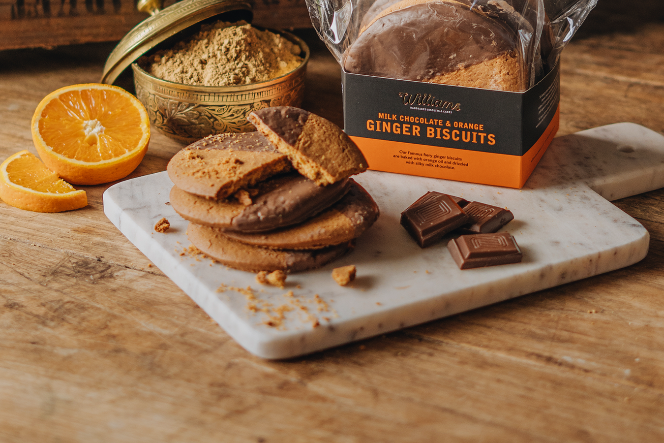 Milk Chocolate and Orange Ginger Biscuits sat on a marble serving board with a sliced orange and ground ginger in the background.