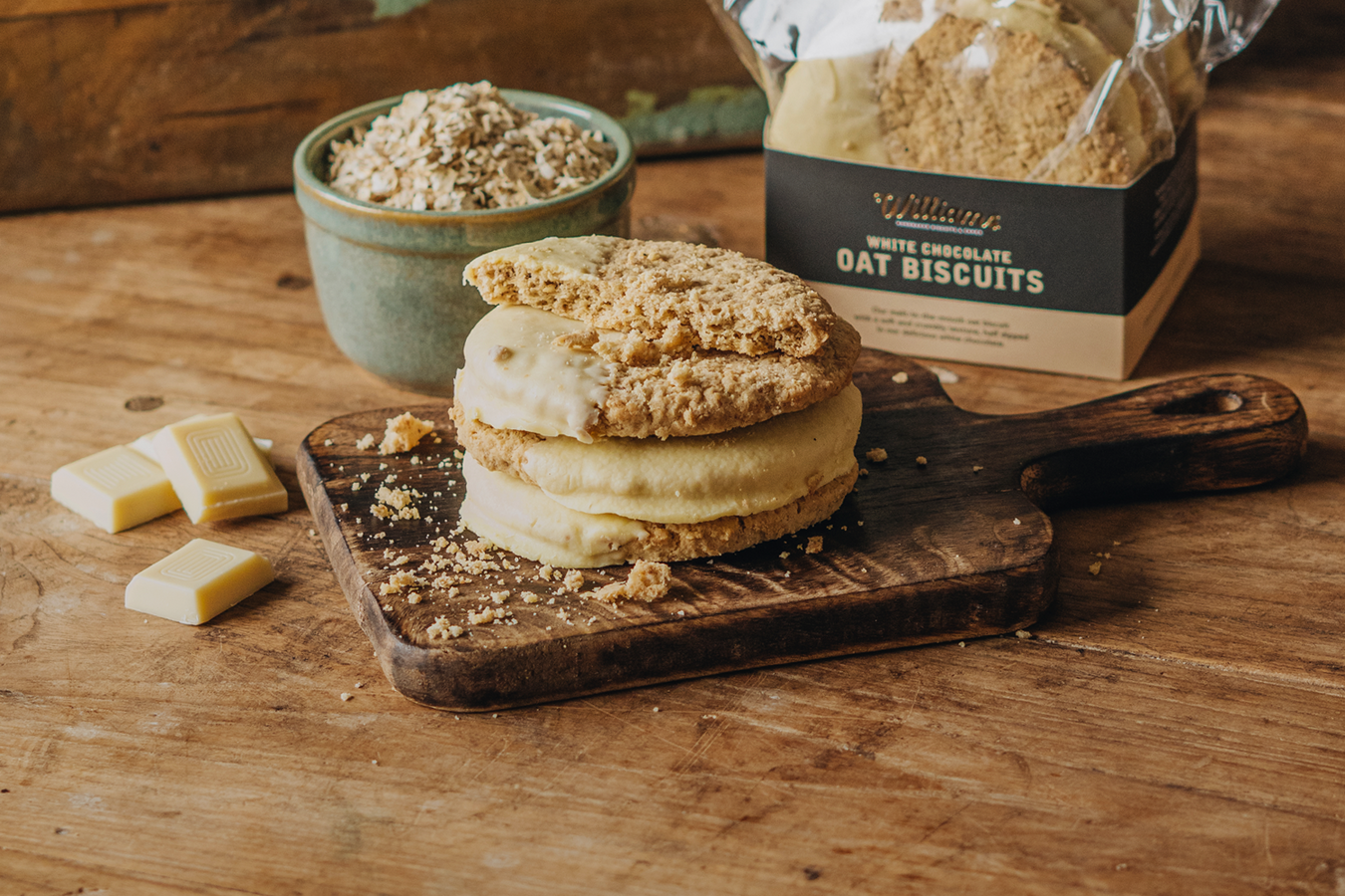 A stack of White Chocolate Oat Biscuits on a wooden block with a packet of biscuits sat behind.
